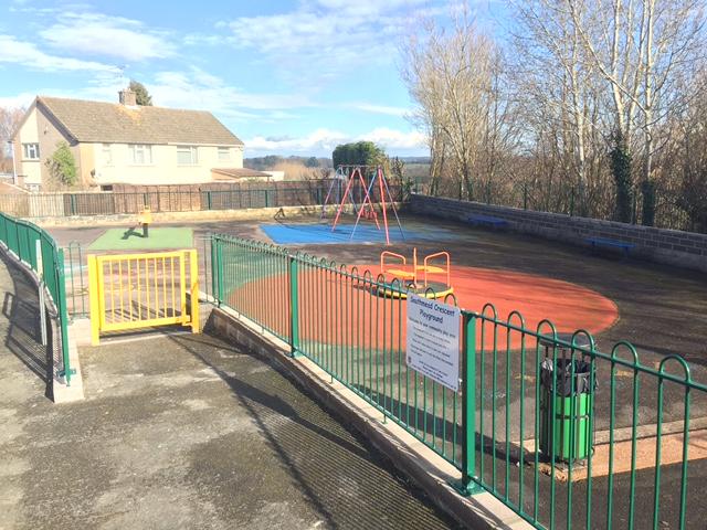 Green railings around children's play area with swings and roundabout
